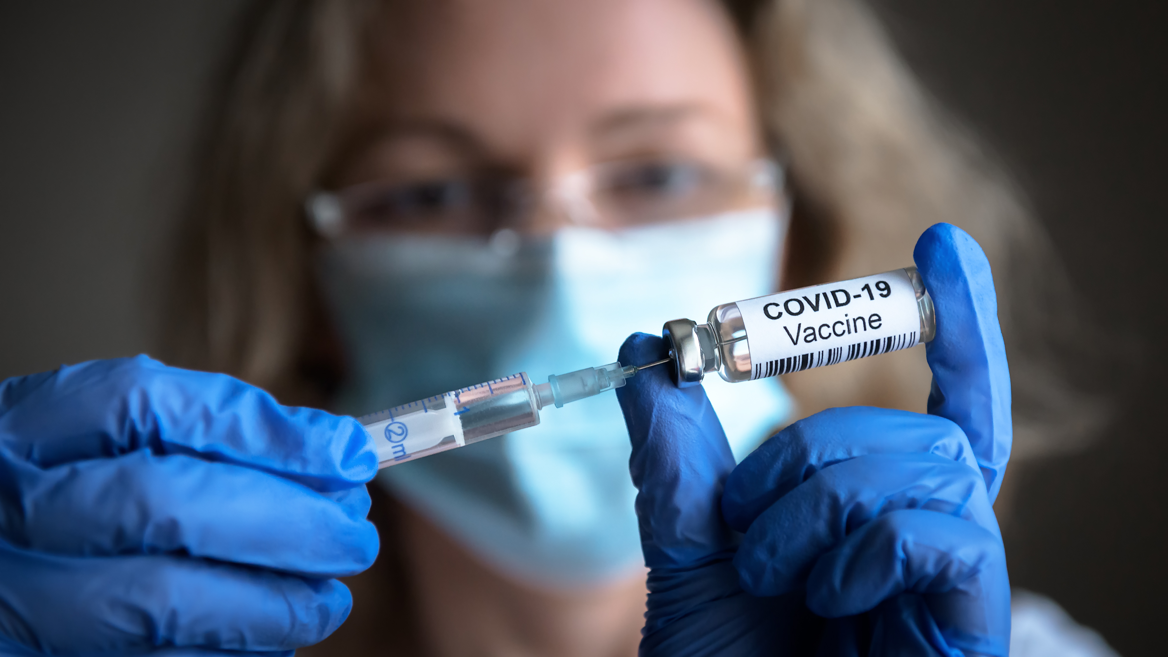 Person med maske trekker opp en vaksinedose mot covid-19 fra et hetteglass. Foto.